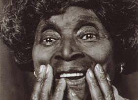 Black and white portrait of Scott smiling, both hands are held to her lower face. Her face and hands fill the frame.